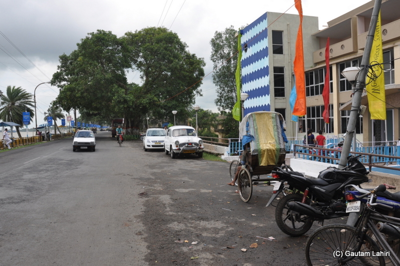 NH 117 became narrow and on the left, we saw the river and on the right the houses, restaurants with cars and bikes loosely parked at Diamond Harbor, West Bengal, India by Gautam Lahiri