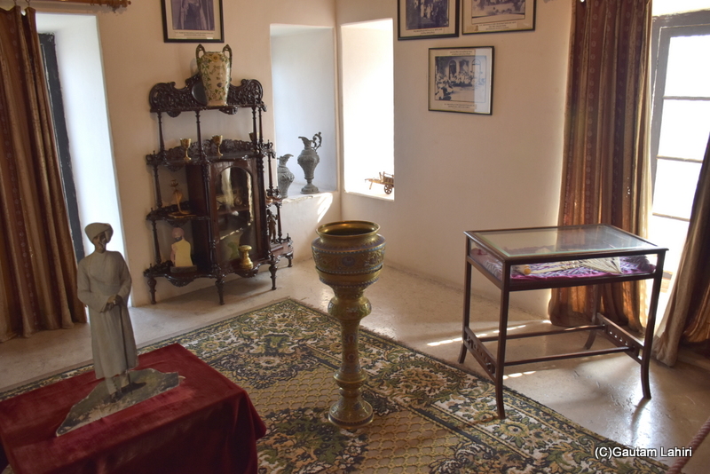 Long drapes hung over the expansive windows, while soothing hues and vintage black-and-white photographs graced the walls. The room was furnished with period pieces, complemented by large, ornate vases. A brass flower vase was prominently displayed on an elegant, sprawling carpet at Naulakha Palace, Gondal. by gautam lahiri