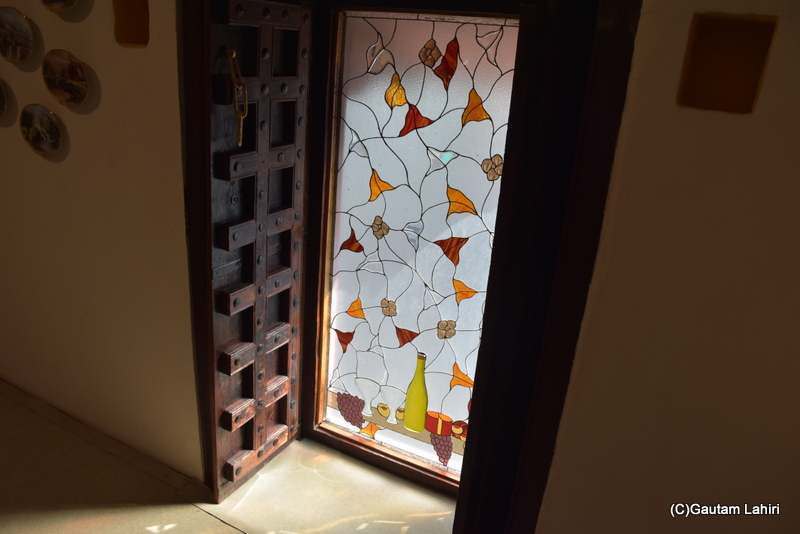 Stained glass adorned with leaf motifs and fruits filters the harsh sunlight partially. The embossed wooden door, its brass chain hanging open, reveals the splendor of Naulakha Palace by gautam lahiri 