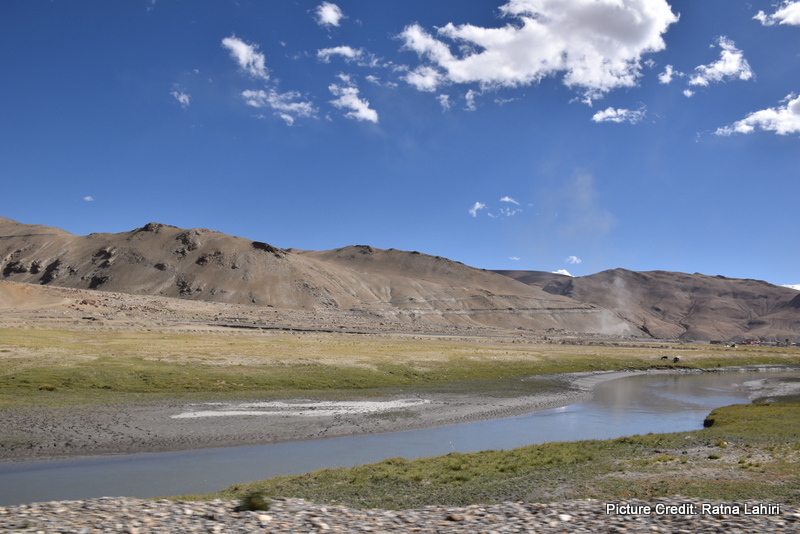 A broken edged shallow river or stream passed by the highway with the grey, brown trans-himalayan range in the distance by gautam lahiri